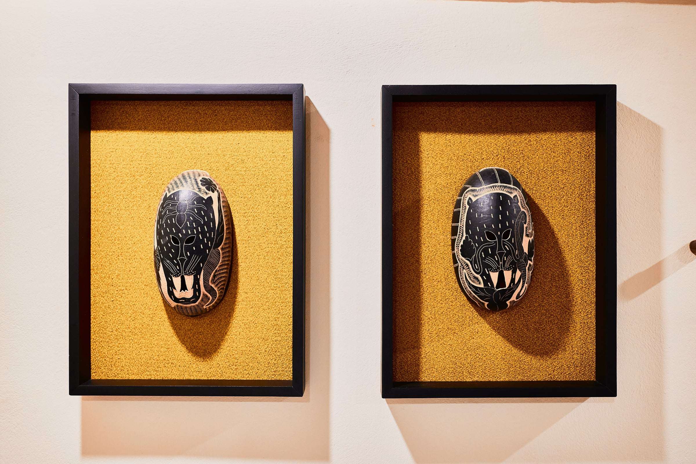 Two hand-carved masks with jaguar faces, in black shadow-box frames on an ochre background