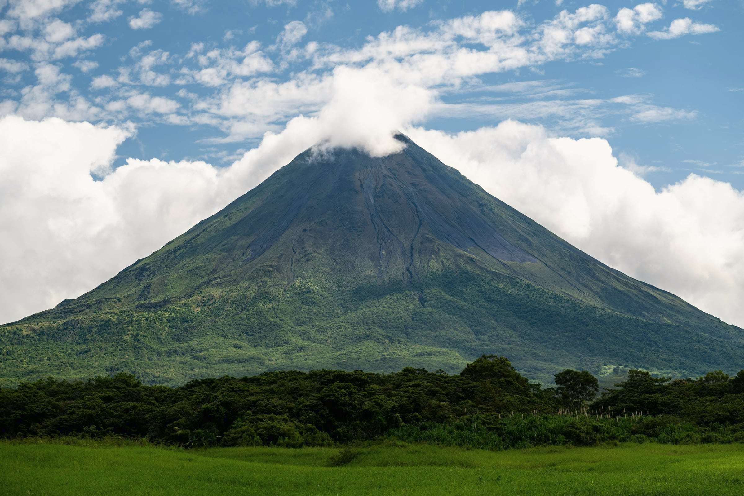 Cone of Arenal Volcano with a cloud on its summit, rising above green forest and pasture near La Fortuna